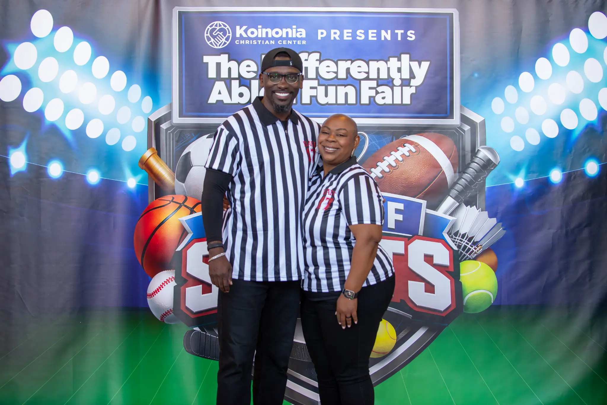 Two smiling attendees in referee shirts in front of the Differently Abled Fun Fair sports-themed backdrop