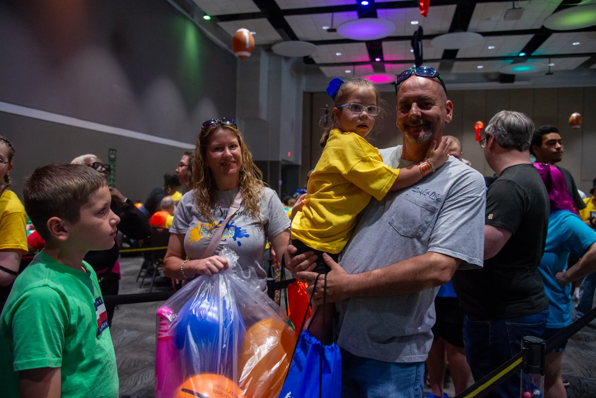 A family enjoying the fair together, with an adult holding a child in a yellow shirt and bags of colorful balloons