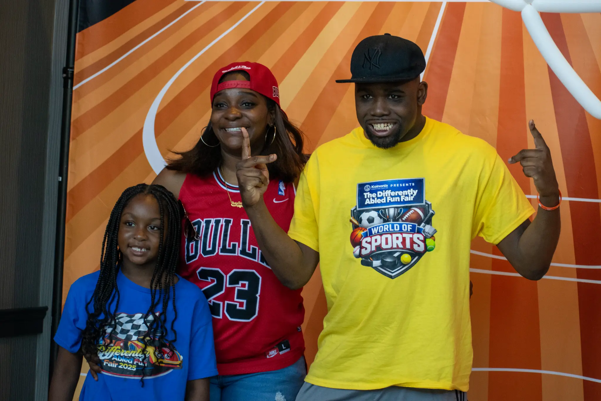 Three smiling fair guests in blue, red, and yellow shirts posing in front of an orange sports-themed backdrop