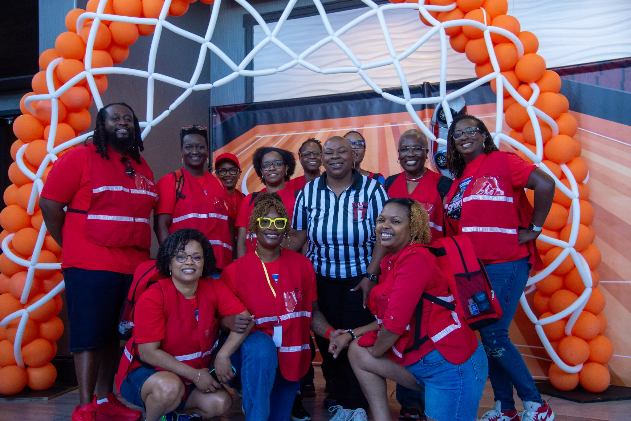 A group in red shirts posing with a referee-shirted attendee beneath an orange basketball balloon arch at DAFF