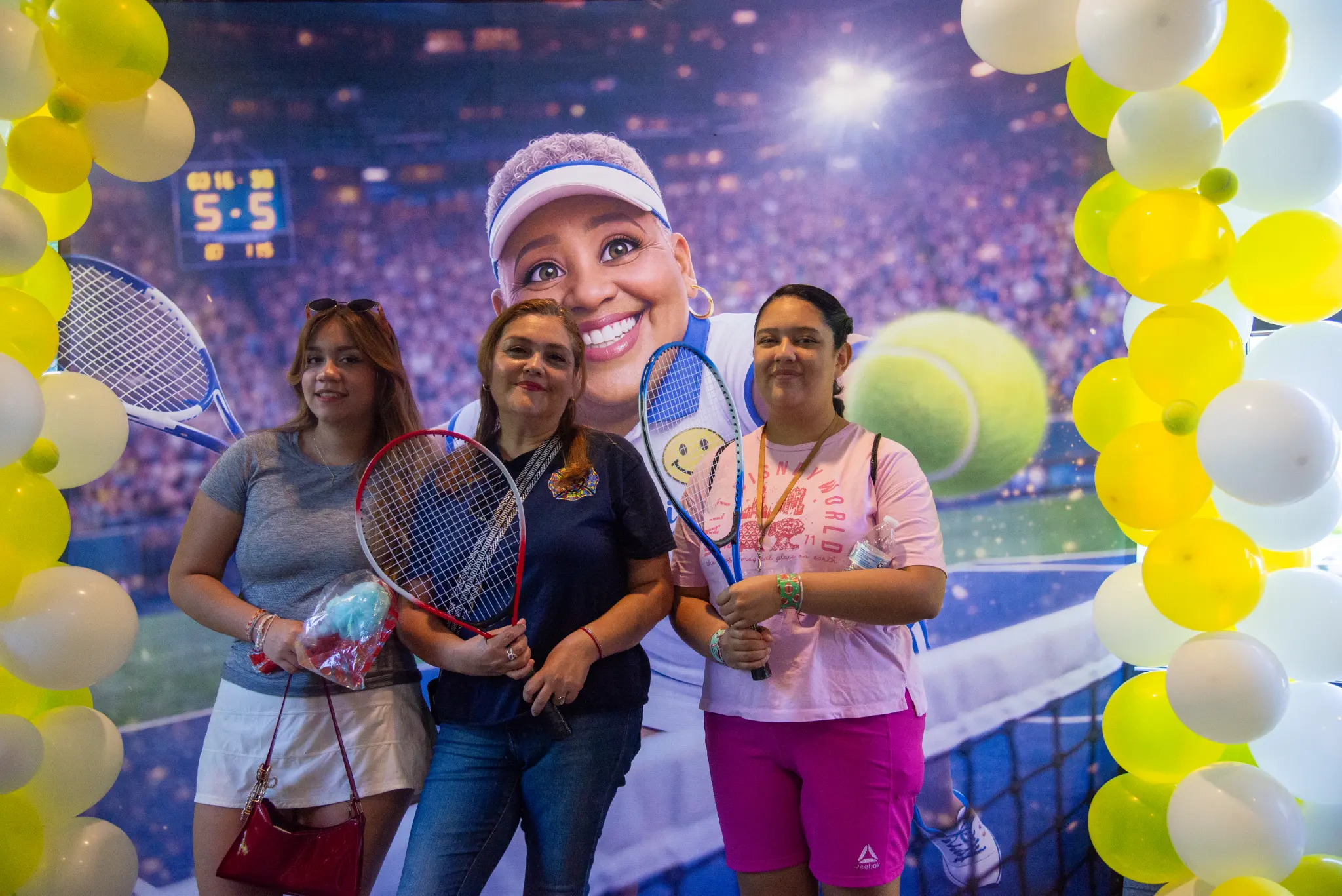 Three fair guests holding tennis rackets in front of a tennis-themed backdrop framed by yellow and white balloons